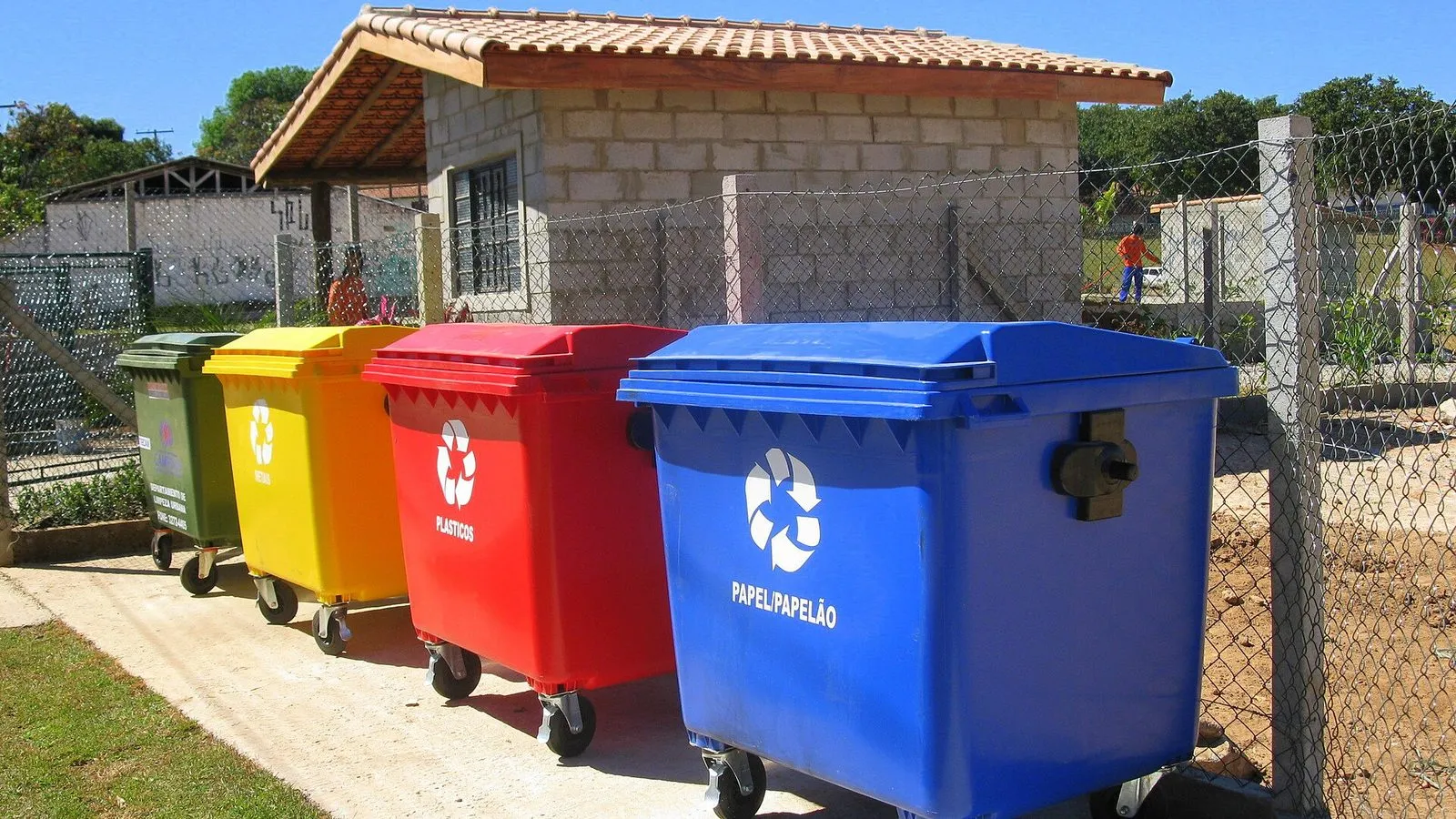 Contêineres coloridos para papel, plástico, metal e vidro em um ecoponto de Campinas, ao lado de uma pequena casa de blocos