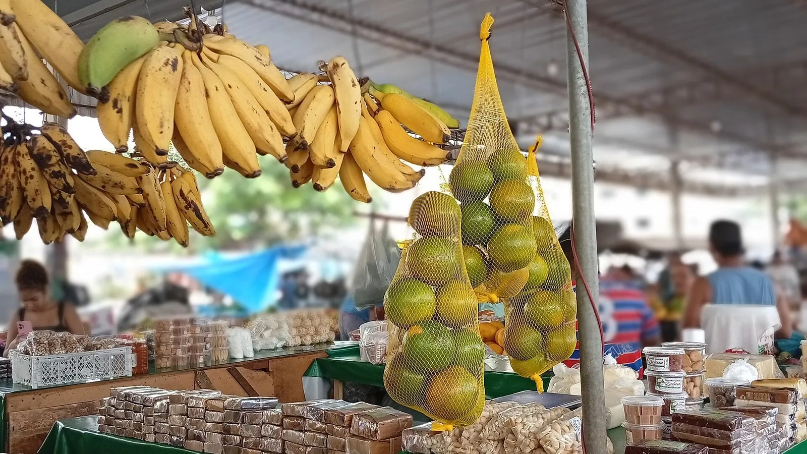 Cachos de banana e sacos de laranja pendurados numa banca de feira livre, com doces e petiscos embalados sobre a mesa