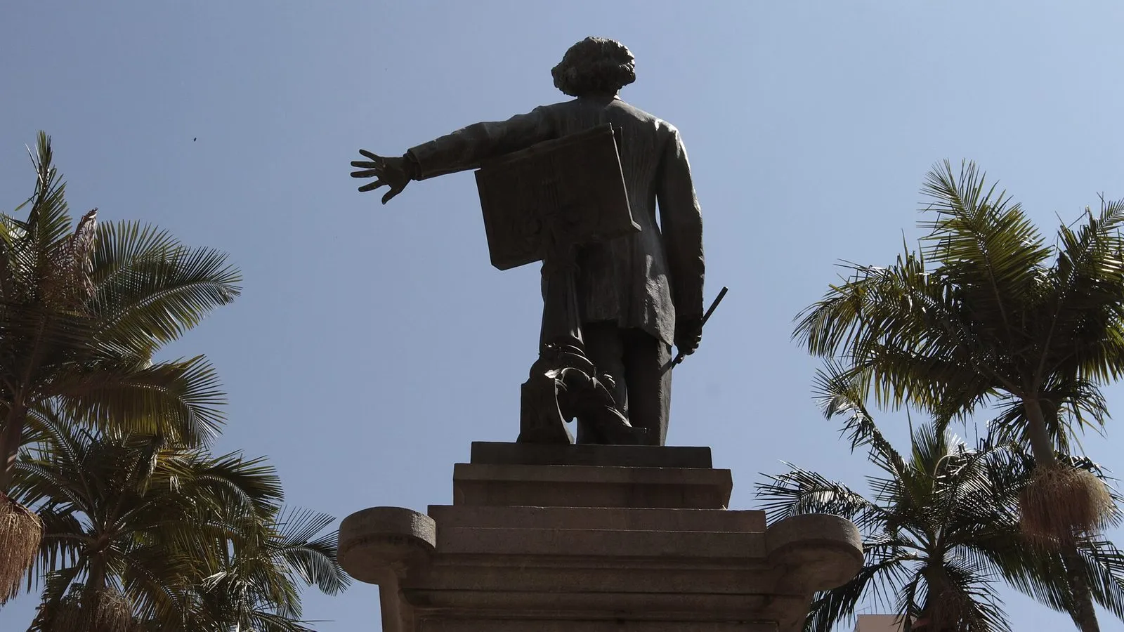 Estátua de bronze de Carlos Gomes no alto do Monumento-Túmulo, vista de costas contra o céu azul, entre palmeiras, no Centro de Campinas