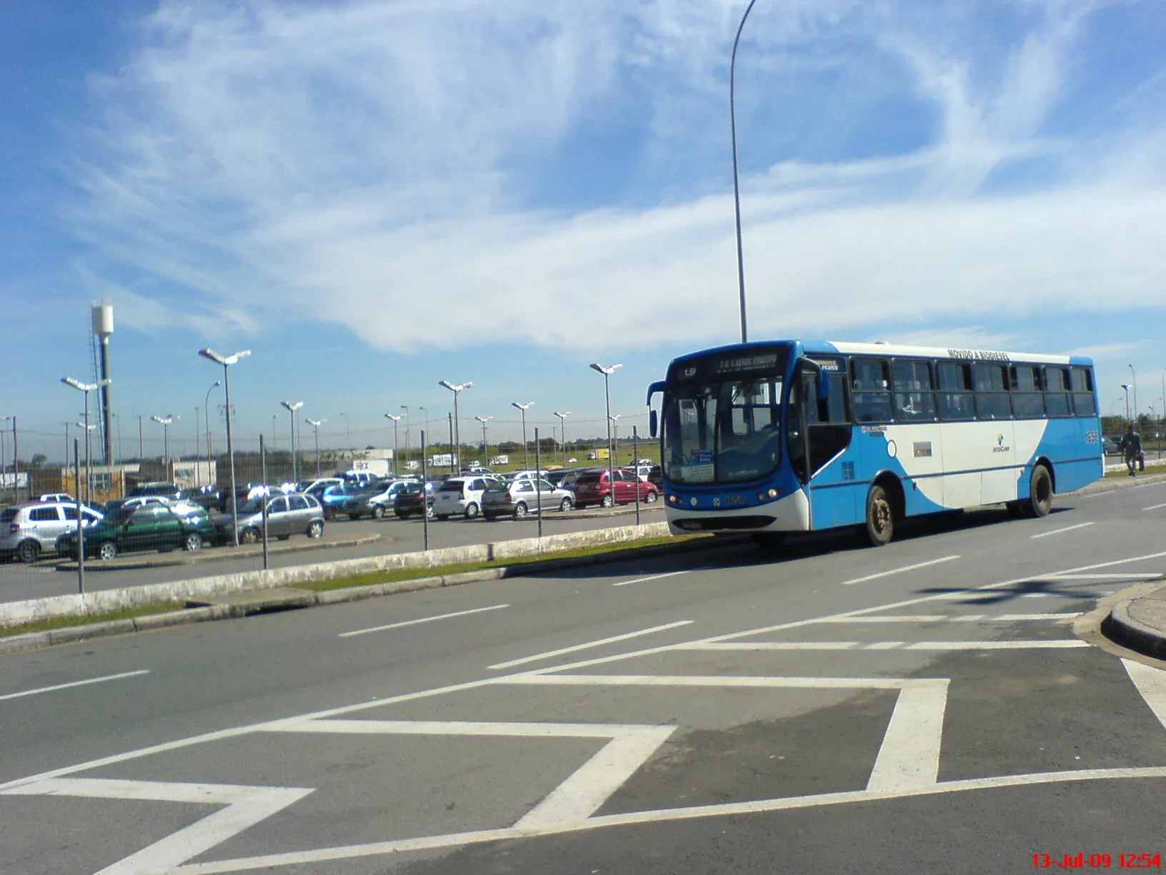 Ônibus urbano azul e branco em via com estacionamento e céu nublado ao fundo