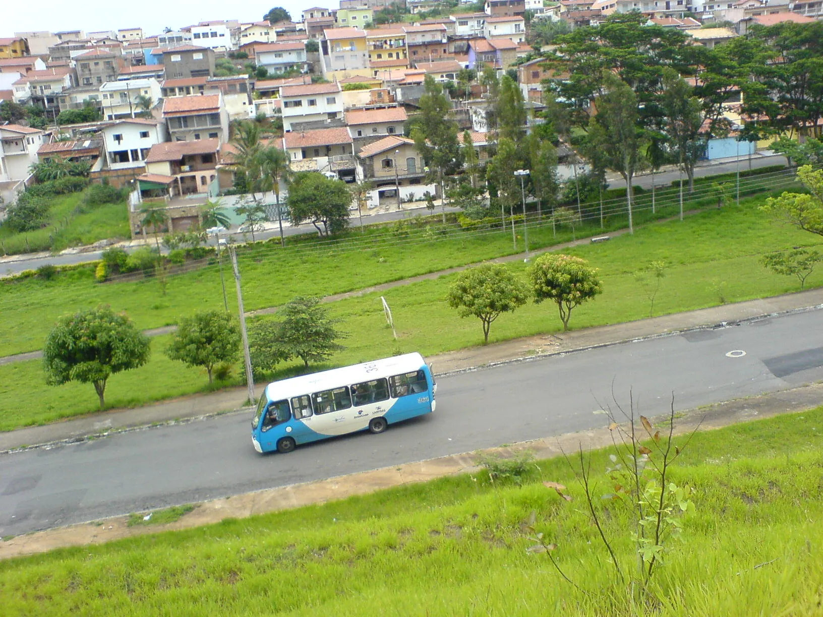 Ônibus urbano azul e branco em via com área verde e casarões ao fundo