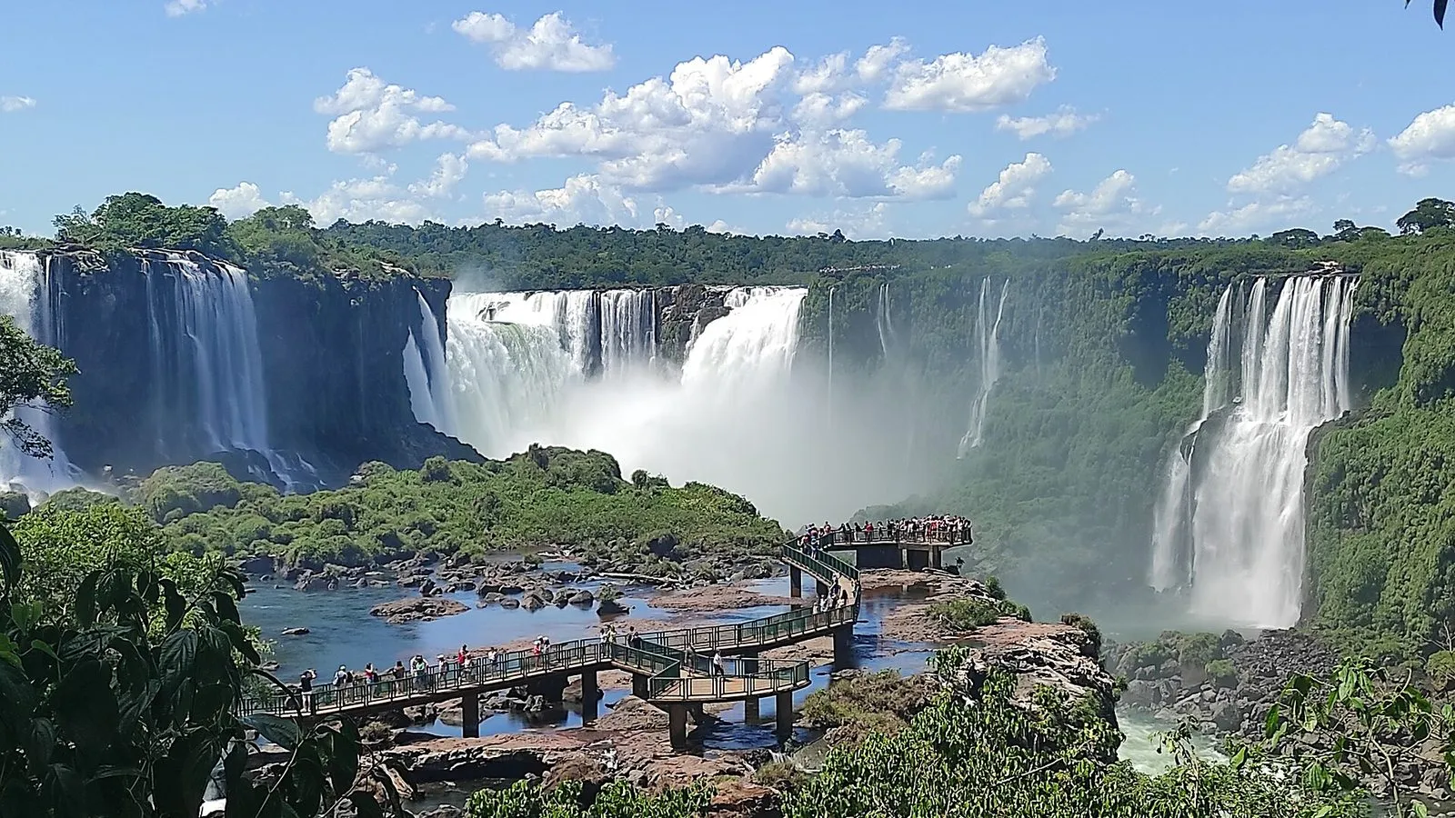 Cataratas do Iguaçu vistas do lado brasileiro, com várias quedas cercadas de mata e a passarela sobre o rio em dia de céu azul