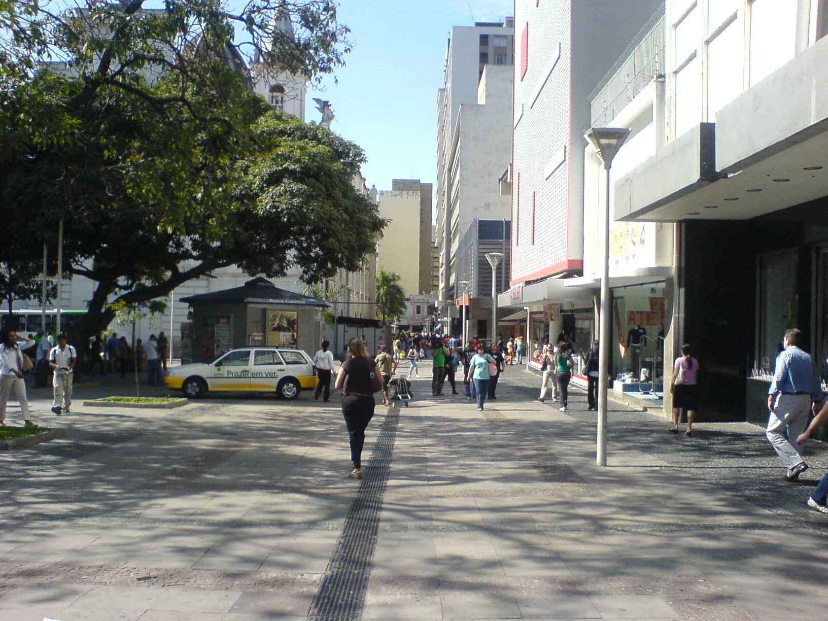 Praça no Centro de Campinas com pessoas circulando, árvore frondosa, lojas e edifícios ao fundo